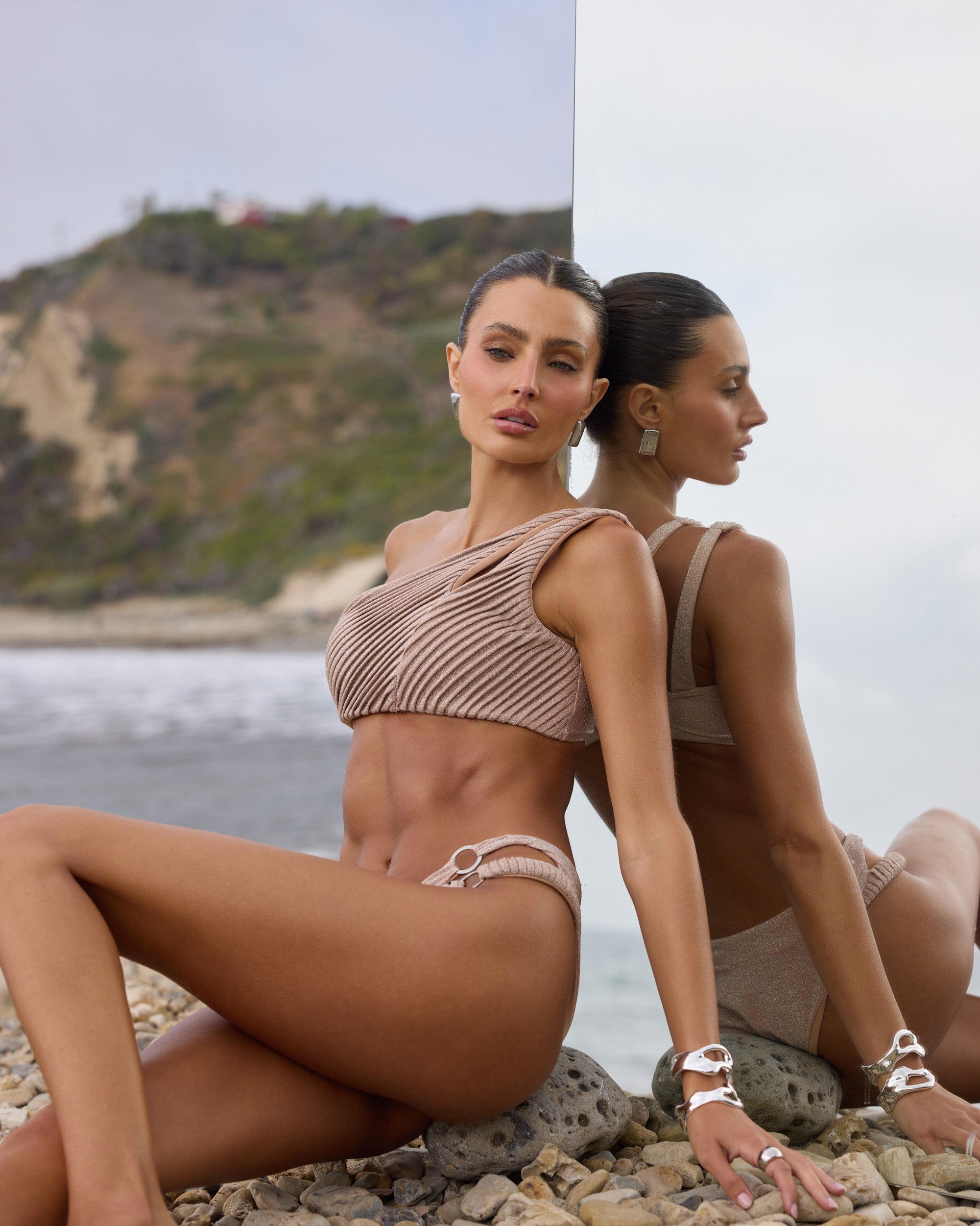 Woman in a beige bikini sits on a rocky beach, leaning against a mirrored surface that reflects her pose. The background shows the ocean and a green hillside under a cloudy sky.
